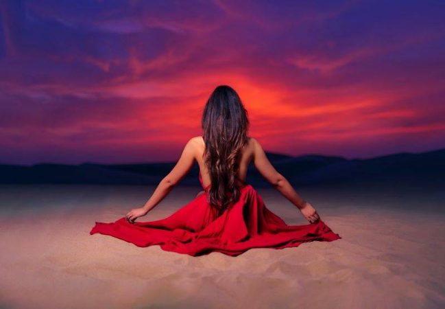 a woman sits looking towards the sunset, with a red dress fanned out around her