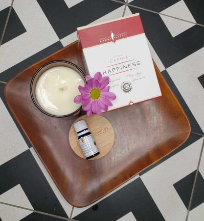 a candle and its box sit on a timber platter next to a bottle of essential oil. The platter sits on a black and white tiled floor