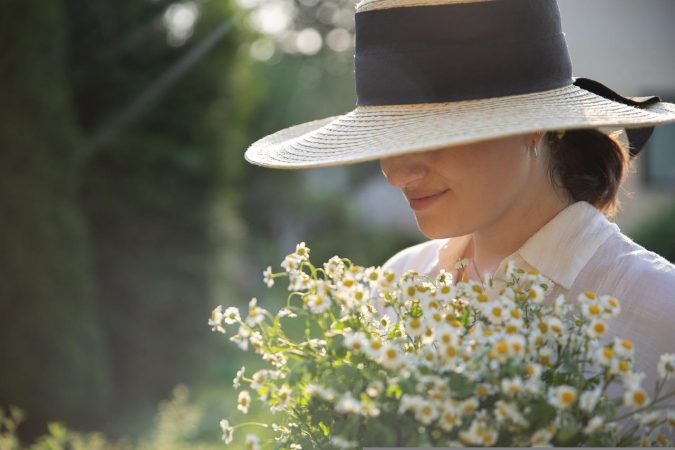 a woman wears a large hat and carries a bunch of daisies