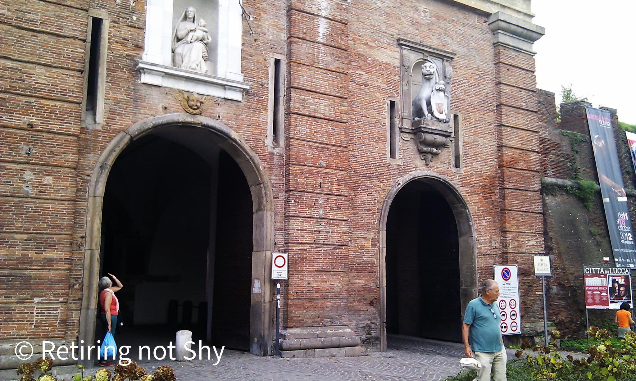 People near the walled entrance to the old City of Lucca, brick walls with arches and statues